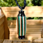 Hearthwood urn pendant with evergreen camo laminate wood grain hanging from a black braided cord in front of a wooden bench with greenery in the background, front-facing view