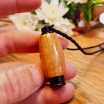 Hearthwood urn pendant with Hawaiian koa wood grain and black stainless steel core held between fingers on a wooden surface with black braided cord and white flowers in the background, close-up view