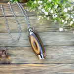 Hearthwood urn pendant with mountain camo wood grain and stainless steel core lying on a rustic wooden surface with silver chain and white flowers, flat lay view