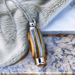 Hearthwood urn pendant with layered multicolor mountain camo wood grain and stainless steel core hanging from a silver chain on fur and stone background, flat lay view