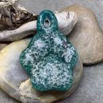 Glass paw print cremation pendant in deep teal with heavy white ash veining, photographed resting on smooth river stones with driftwood in the background