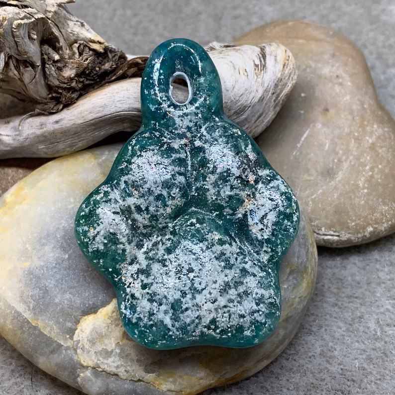 Glass paw print cremation pendant in deep teal with heavy white ash veining, photographed resting on smooth river stones with driftwood in the background
