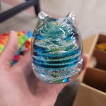 Hand holding a glass cat-shaped memorial paperweight featuring layered swirls of blue and teal color mixed with cremation ash suspended inside clear glass, with subtle reflections and a rounded sculpted body