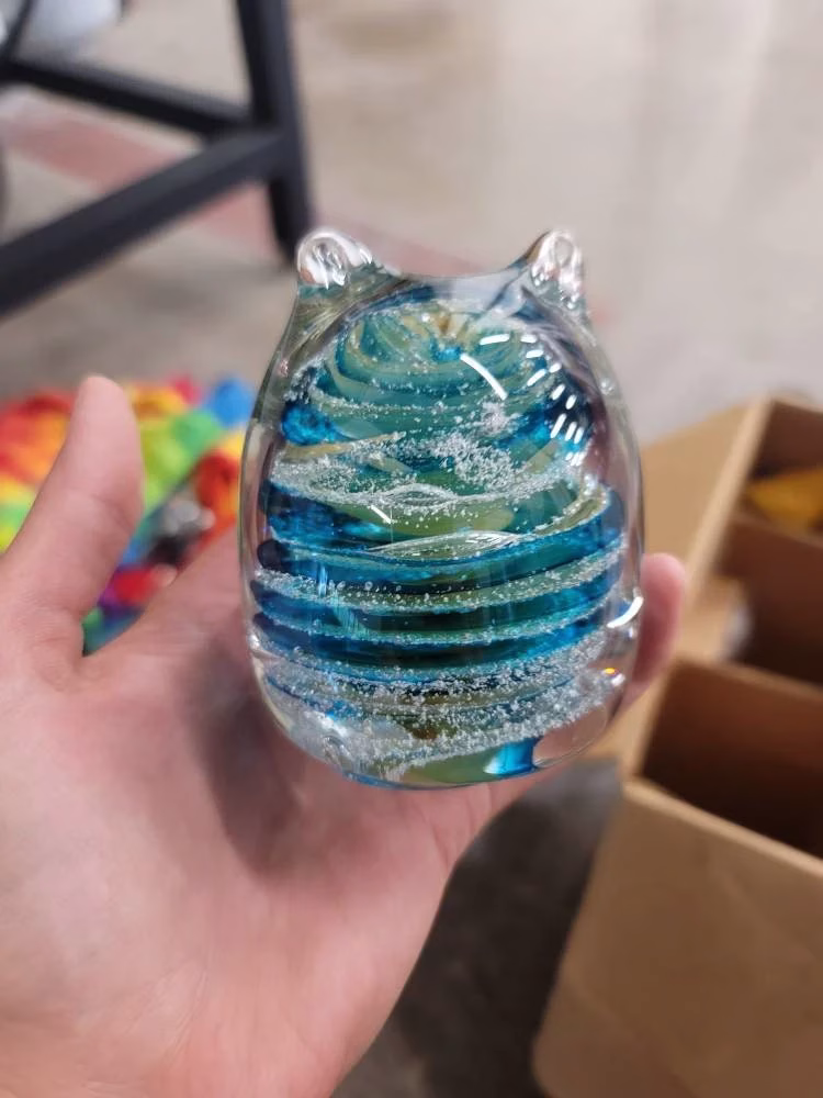 Hand holding a glass cat-shaped memorial paperweight featuring layered swirls of blue and teal color mixed with cremation ash suspended inside clear glass, with subtle reflections and a rounded sculpted body