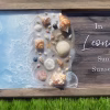 Oceanside memorial artwork featuring a resin shoreline with seashells, starfish, and cremation ash details blending into soft blue ocean waves beside a wooden panel with engraved memorial text.