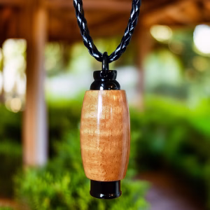 Hearthwood urn pendant with Hawaiian koa wood grain and black stainless steel core hanging from a black braided cord outdoors with greenery and wooden structure in the background, front-facing view