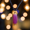 Hearthwood urn pendant with pink camo layered wood grain and gold stainless steel core hanging from a black braided cord in front of warm blurred string lights, front-facing view