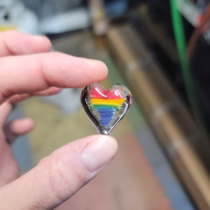 Hand holding a small clear glass heart-shaped cremation stone featuring layered rainbow colors—red, orange, yellow, green, blue, and purple—encased inside, with subtle ash patterns suspended within the glass.