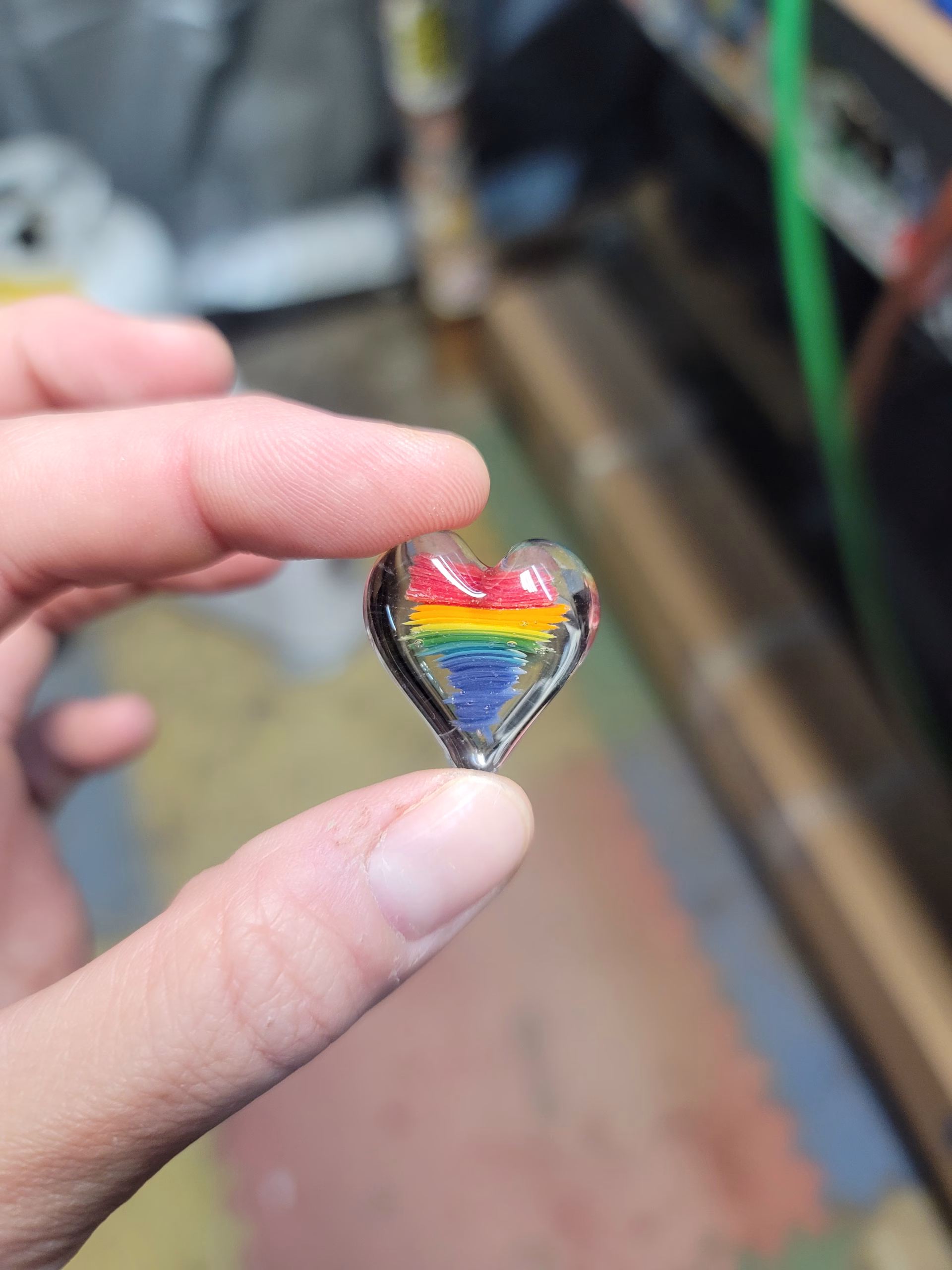 Hand holding a small clear glass heart-shaped cremation stone featuring layered rainbow colors—red, orange, yellow, green, blue, and purple—encased inside, with subtle ash patterns suspended within the glass.