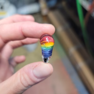 Side view of a clear glass heart-shaped cremation stone held between fingers, showing layered rainbow colors—red through purple—encased inside the glass with subtle cremation ash patterns.