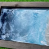 Rectangular Oceanside memorial artwork featuring swirling blue and white resin waves blended with cremation ash, set in a wooden frame with seashells and a small starfish along the sandy edge.