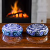 Two blue and aqua glass cremation stones with spiral cores and pinwheel-edged patterns resting on a polished wooden surface with a glowing fireplace in the background
