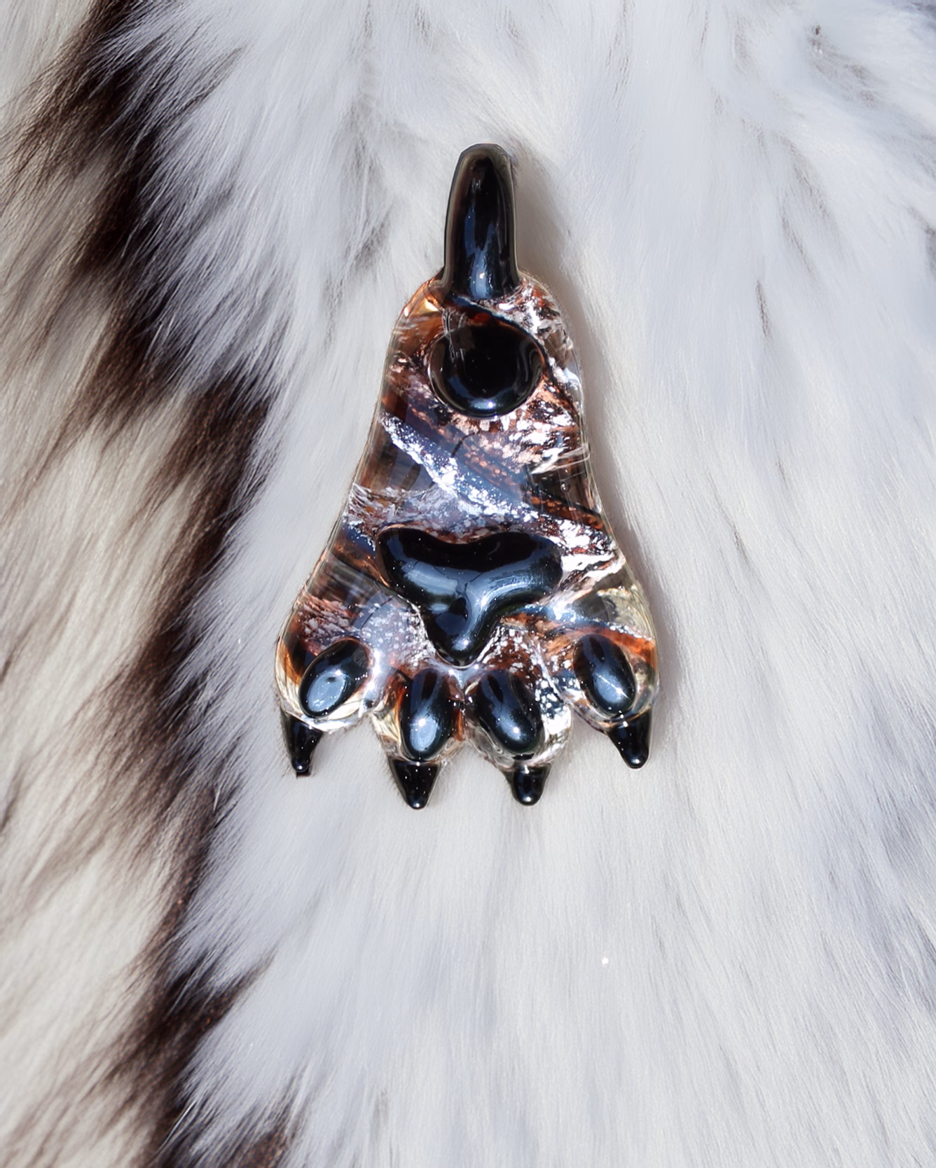 Glass paw cremation pendant with black paw pads and copper and silver swirled glass resting on a white fur background