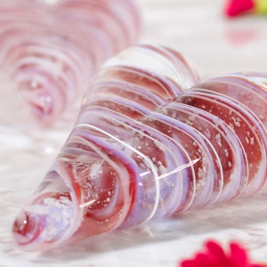 Close-up of a translucent pink and purple glass love heart cremation paperweight with visible cremation ash, resting on a marble surface with scattered rose petals