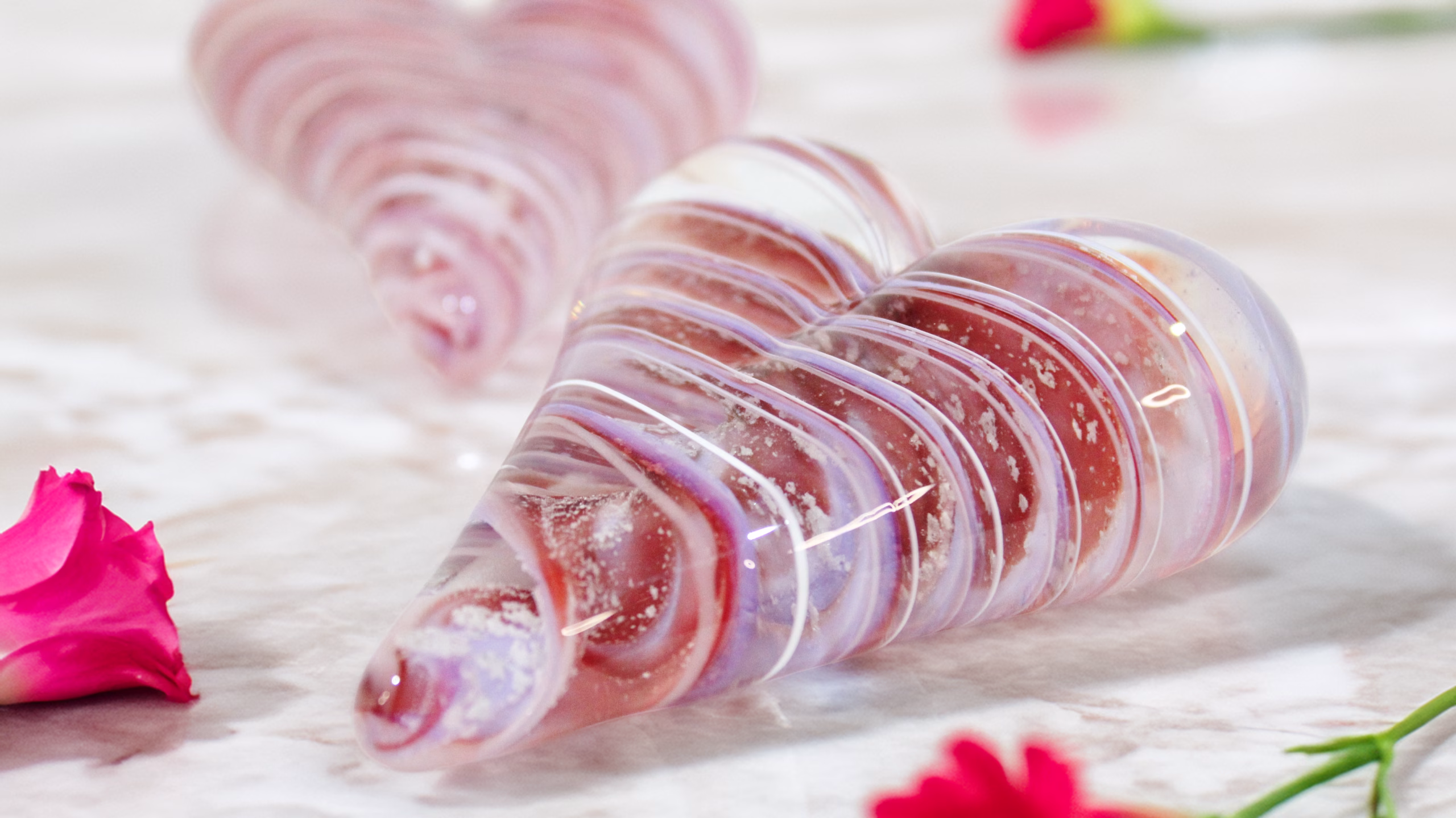 Close-up of a translucent pink and purple glass love heart cremation paperweight with visible cremation ash, resting on a marble surface with scattered rose petals