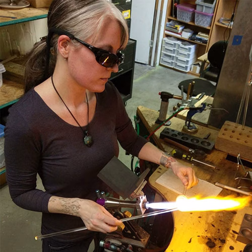 Glass artist working at a torch in a jewelry studio while shaping molten glass with hand tools during the glassblowing process
