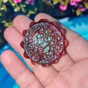 Front view of a deep red sensory cremation stone with embedded cremation ash, shimmering speckled texture, and scalloped glass edging, resting on an open palm in natural sunlight