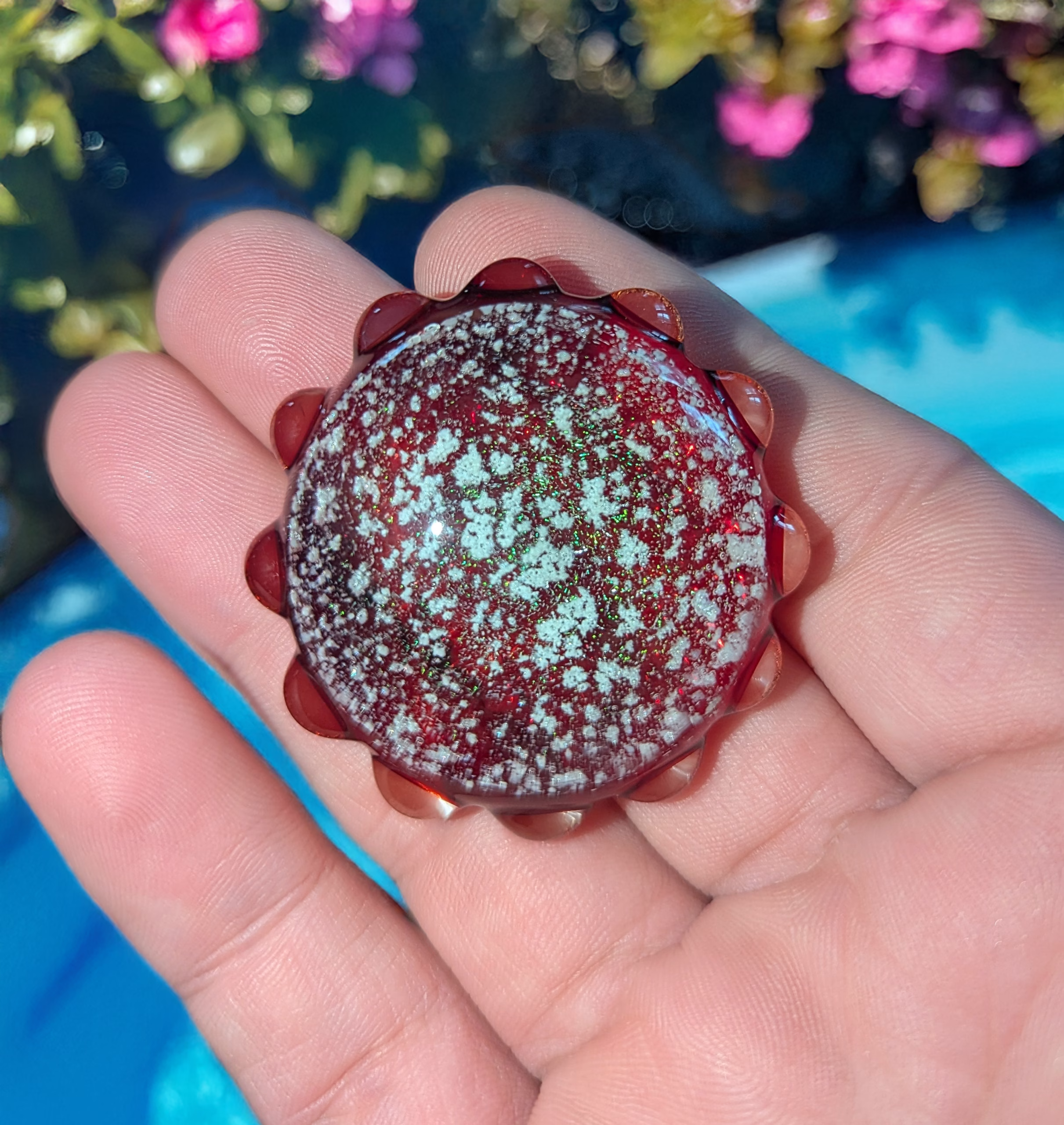 Front view of a deep red sensory cremation stone with embedded cremation ash, shimmering speckled texture, and scalloped glass edging, resting on an open palm in natural sunlight