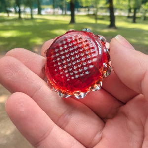 Backside view of a deep red sensory cremation stone with raised bubble texture and stamped pattern, held between fingers, showing smooth glass edges and dimensional surface detail