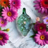 Teardrop glass cremation pendant with green transparent body, suspended opal center, and subtle ash infusion, photographed on white marble surrounded by pink and purple flowers
