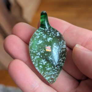Opal teardrop glass cremation pendant with ashes in green glass, featuring suspended cremation ash and a glowing opal accent, handcrafted memorial jewelry held in hand for scale