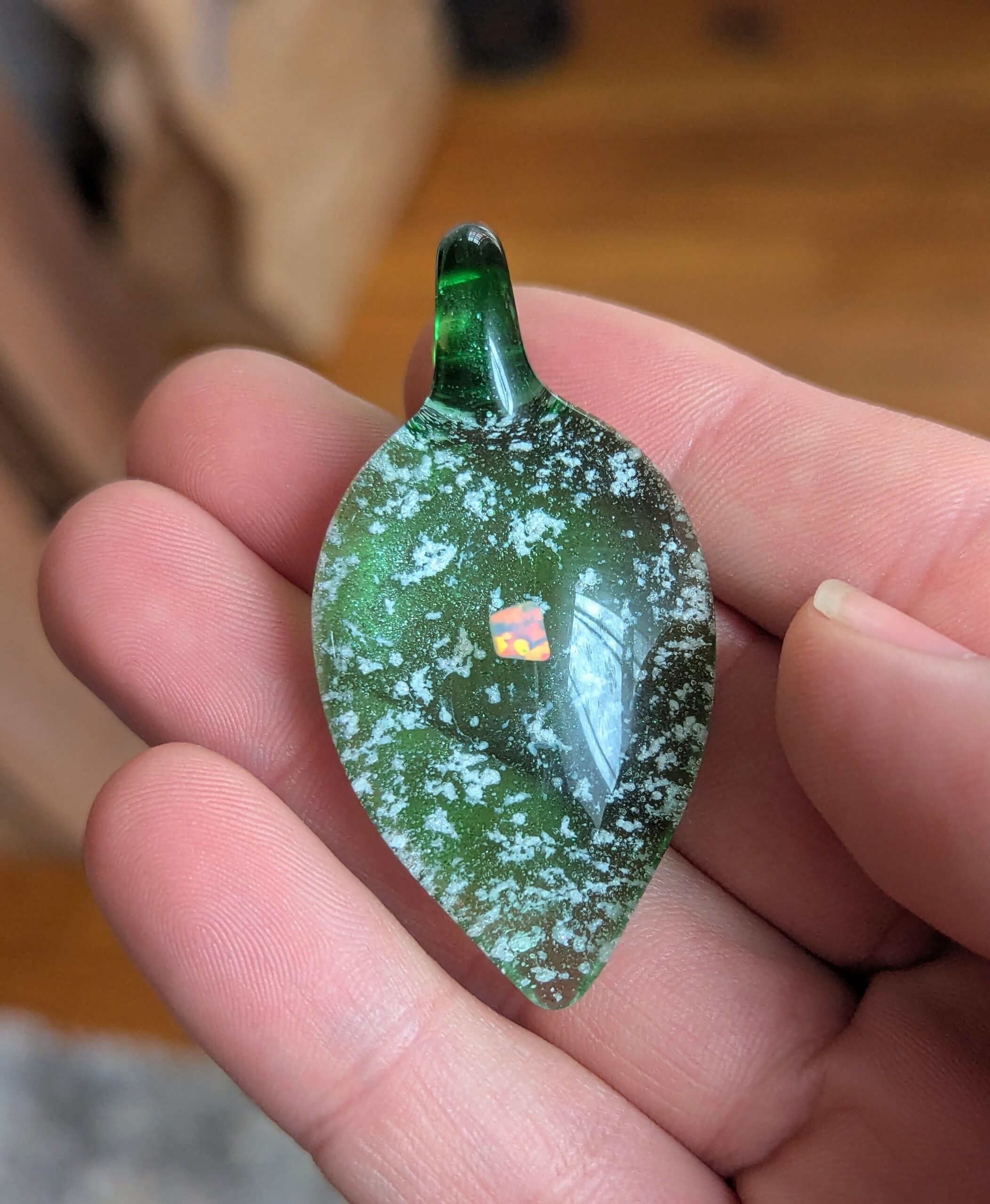 Opal teardrop glass cremation pendant with ashes in green glass, featuring suspended cremation ash and a glowing opal accent, handcrafted memorial jewelry held in hand for scale