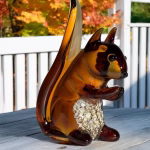 Amber glass squirrel infused with cremation ash sitting on a wooden deck with white railing and autumn trees in the background