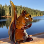 Amber glass squirrel infused with cremation ash sitting on a wooden railing overlooking a lake and forest
