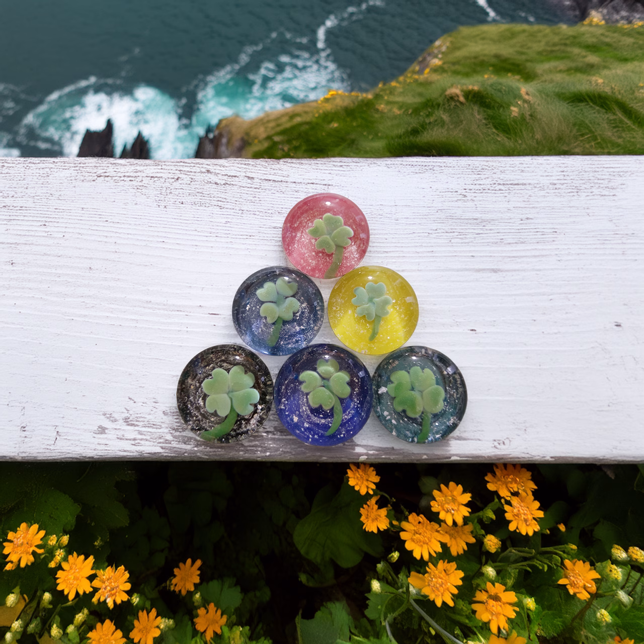 Six round glass four-leaf clover cremation stones in various colors arranged on a white wooden ledge with ocean cliffs and yellow flowers in the background