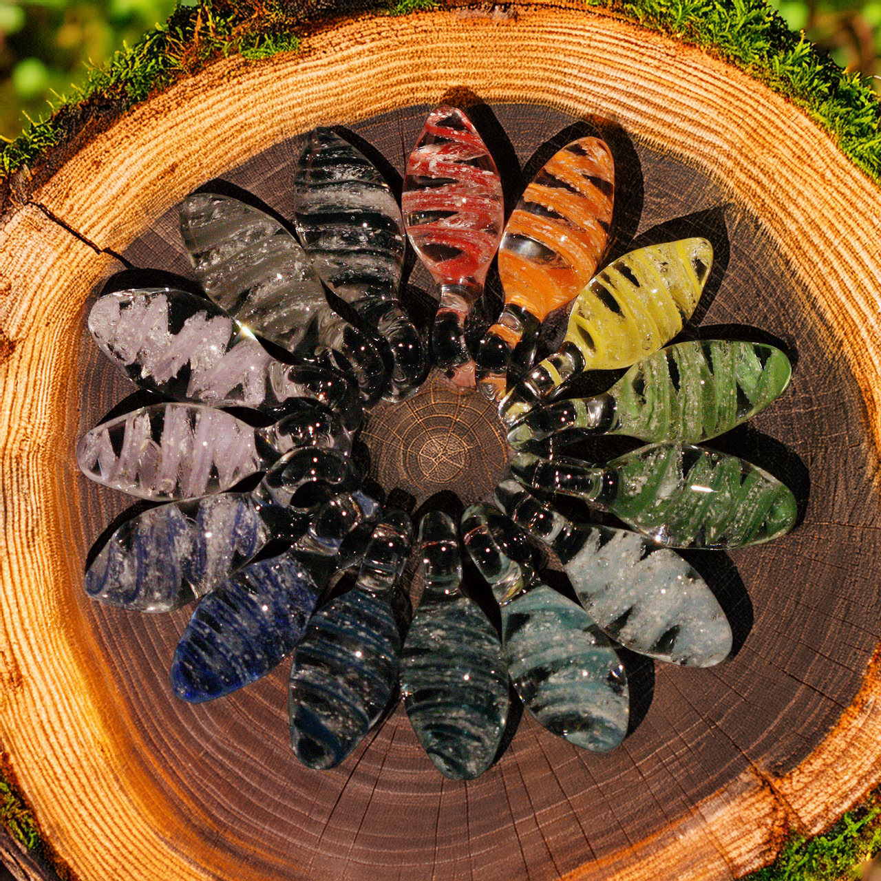 Assorted spiral drop cremation pendants in multiple colors arranged in a circular pattern on a natural wood slice with moss edging
