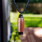 Hearthwood urn pendant in black walnut and maple wood with black stainless steel core hanging from a braided cord outdoors with blurred green yard and wooden deck background
