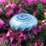Blue and aqua glass cremation stone with spiral core and pinwheel-edged pattern resting on vibrant pink flowers