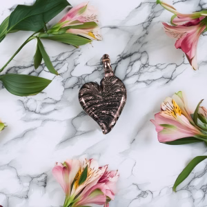 Black glass ribbon heart cremation pendant with soft rose tint and silver ribbon swirl detail on white marble background with pink flowers