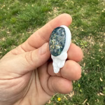Angled side view of the Stealie cremation pendant showing the blue moon bloom glass front with visible gold flecks and the white glass skull profile, held between fingers outdoors against green grass