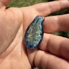 Teardrop-shaped glass cremation pendant with translucent blue edges and gold petal-like pattern cradled in the palm of a hand outdoors in sunlight