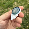 Front angled view of the Stealie cremation pendant showing the blue moon bloom glass design with visible gold ash flecks set in a white glass skull, held between fingers outdoors against green grass