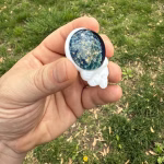 Angled side view of the Stealie cremation pendant showing the blue moon bloom glass front with visible gold flecks and the white glass skull profile, held between fingers outdoors against green grass