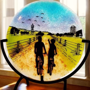 Countryside Bike Ride Suncatcher with Cremation Ash showing two cyclists riding down a fenced country road at sunset with cremation ash blended into the blue and purple sky