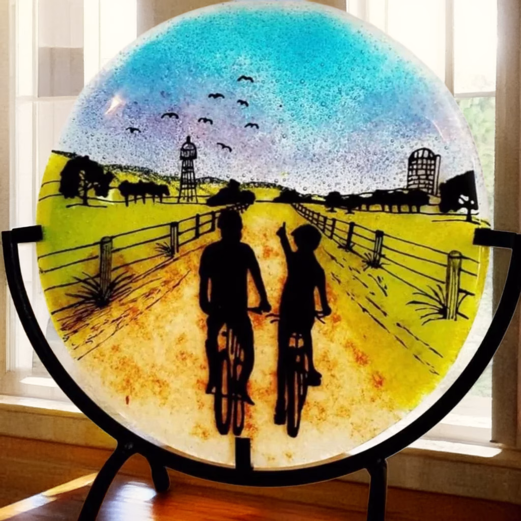 Countryside Bike Ride Suncatcher with Cremation Ash showing two cyclists riding down a fenced country road at sunset with cremation ash blended into the blue and purple sky