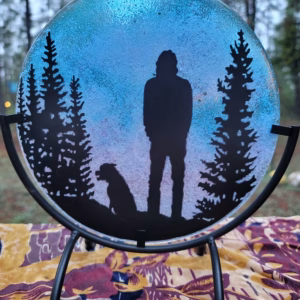 Forest Pines Suncatcher with Cremation Ash displayed on a metal stand outdoors, showing a silhouetted person and dog framed by pine trees against a blue sky