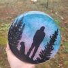 Forest Pines Suncatcher with Cremation Ash shown handheld outdoors with a standing figure and a seated dog silhouetted between tall pine trees against a blue and pink sky with cremation ash blended into the sky