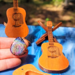 Functional Glass Guitar Pick with Cremation Ash in Irid held in front of wooden guitar-shaped display stand and matching wooden guitar display box outdoors