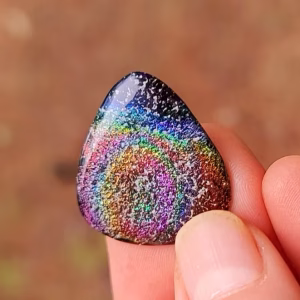 Functional Glass Guitar Pick with Cremation Ash in Irid held between fingers, showing a rainbow iridescent swirl pattern across the glass surface