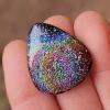 Functional Glass Guitar Pick with Cremation Ash in Irid held between fingers, showing a rainbow iridescent swirl pattern across the glass surface