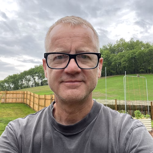 Front-facing outdoor portrait of artist Paul K wearing black glasses and a gray shirt, standing in a grassy backyard with garden beds, fencing, and cloudy sky in the background