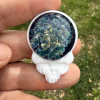 Stealie-shaped glass cremation pendant with blue moon floral design and white skull-style body held between fingers outdoors against green grass