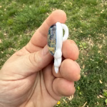 Side profile of Stealie cremation pendant showing the white glass skull body and looped bail with the blue moon bloom glass center visible, held between fingers outdoors over green grass