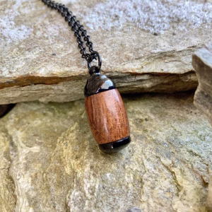 Angled top-down view of an extra small Hearthwood urn pendant made from Hawaiian Koa wood with polished black metal end caps, resting on a stone surface and attached to a black chain for holding cremation ashes.