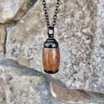 Straight-on view of an extra small Hearthwood urn pendant made from Hawaiian Koa wood with polished black metal end caps, suspended from a black chain for holding cremation ashes.