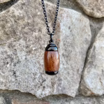 Straight-on view of an extra small Hearthwood urn pendant made from Hawaiian Koa wood with polished black metal end caps, suspended from a black chain for holding cremation ashes.
